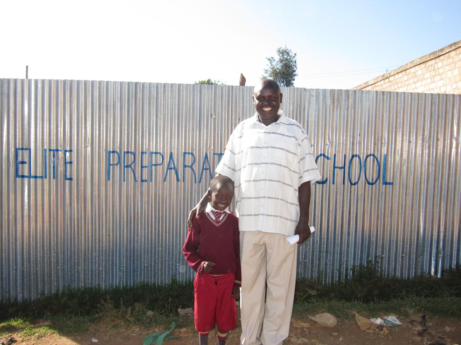 Simeon, a tailor in a small town in Kenya, with his youngest son smiling together proudly outside the school his son attends. Simeon pays his for all of his children's school fees with the profits from his tailoring business which he has expanded with Kiva loans. Jada, Kenya