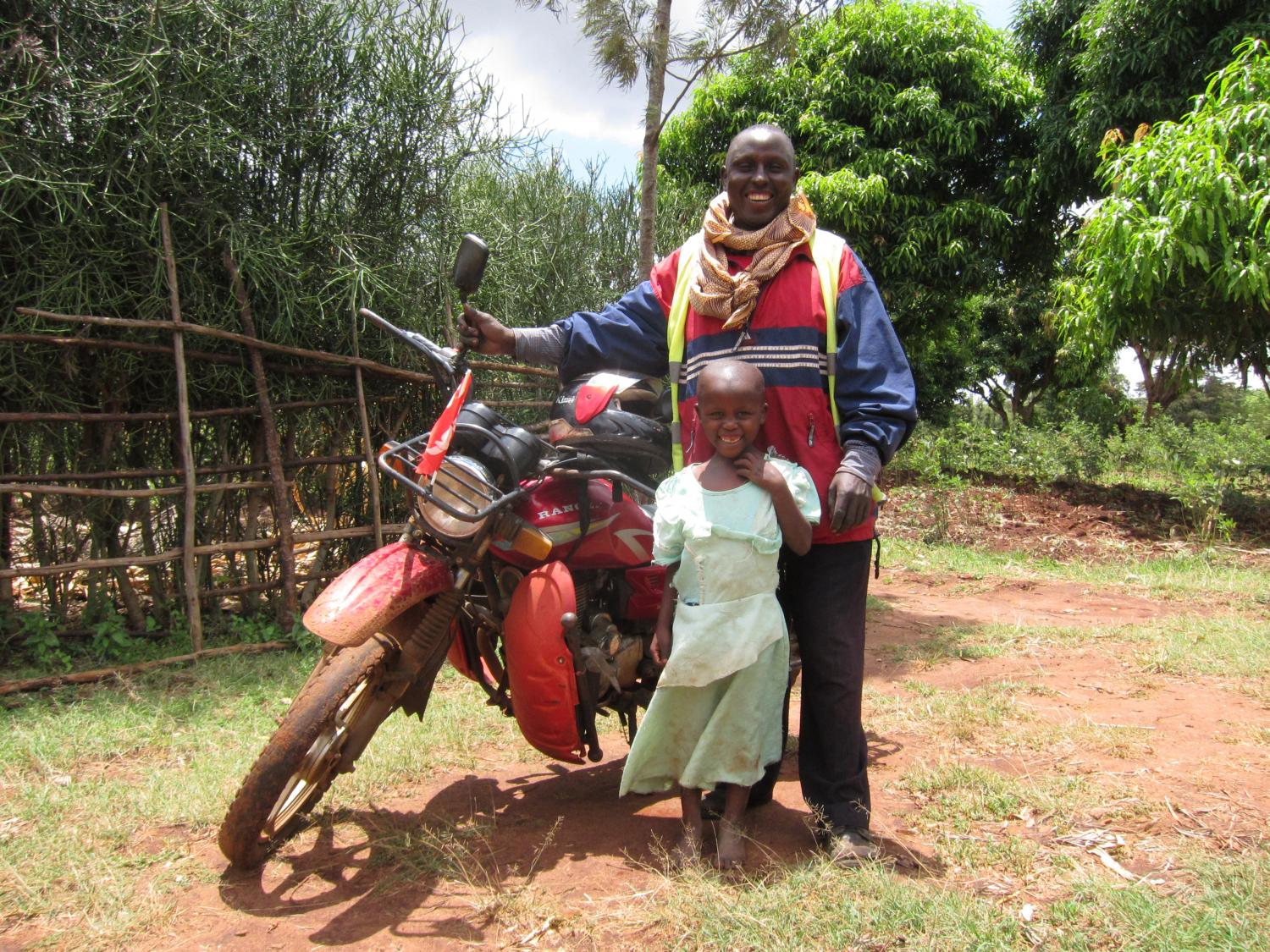 Nicholas and his daughter smiling with his boda boda at his home.