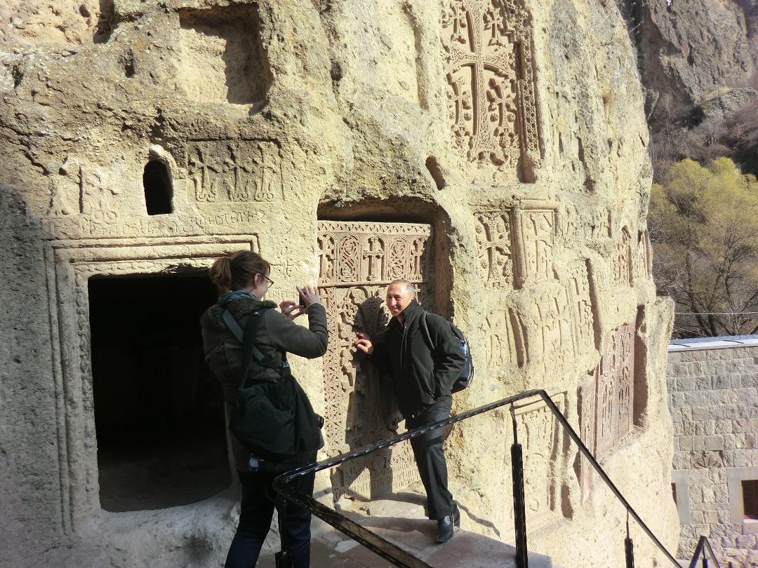 Liz photographs Gagik by carved blooming crosses on cave's outer wall at Gerhard